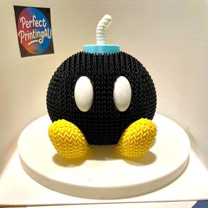 May include: A 3D-printed black Bob-omb character with white eyes, yellow feet, a light blue fuse base, and a white fuse. The character is sitting on a white circular base. The text "Perfect PrintingAU" is visible.