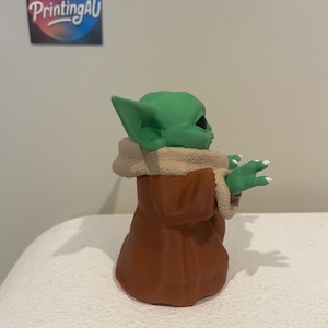 May include: A green figurine of a character with large ears, wearing a brown robe with a tan fur-like collar. The figurine is on a white surface. The text "Perfect PrintingAU" is on a sticker in the background.