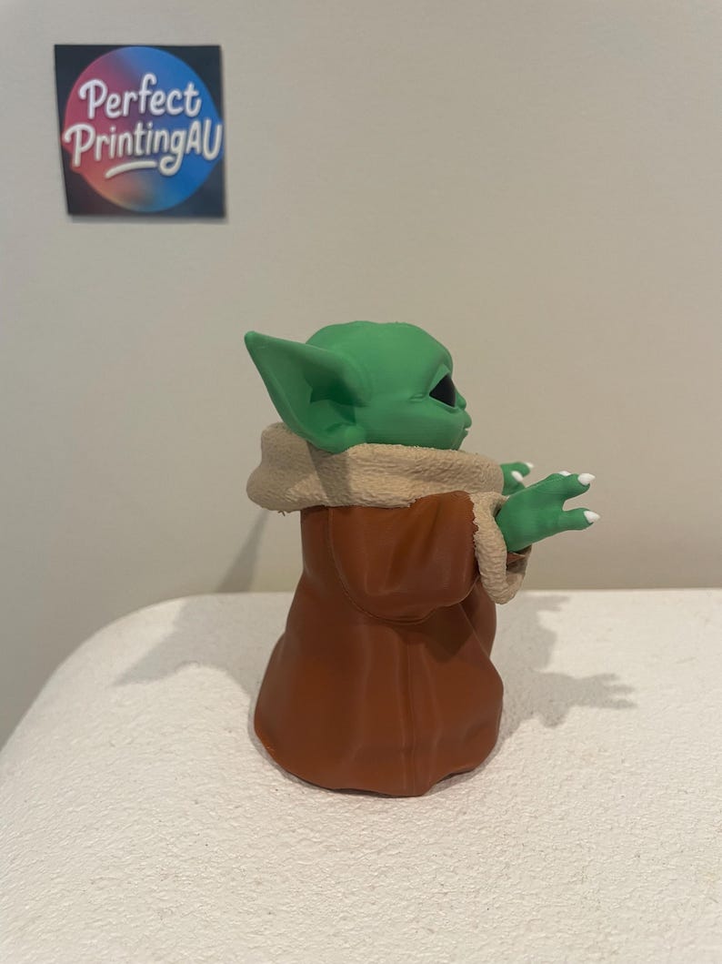 May include: A green figurine of a character with large ears, wearing a brown robe with a tan fur-like collar. The figurine is on a white surface. The text "Perfect PrintingAU" is on a sticker in the background.