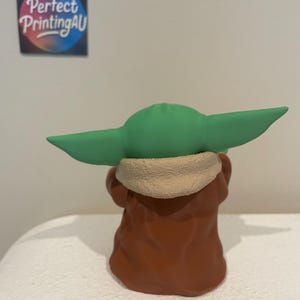 May include: A figurine of a character with large green ears, wearing a brown robe with a tan collar. The figurine is viewed from the back. A sticker with the text "Perfect Printing AU" is visible in the background.