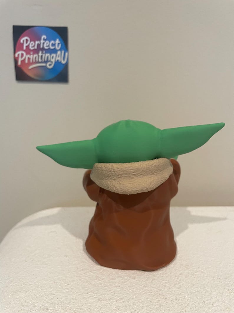 May include: A figurine of a character with large green ears, wearing a brown robe with a tan collar. The figurine is viewed from the back. A sticker with the text "Perfect Printing AU" is visible in the background.