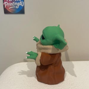 May include: A green figurine of a character with large ears and a brown robe. The figurine has white fingernails and is standing on a white surface. The figurine is a character from a popular science fiction franchise.