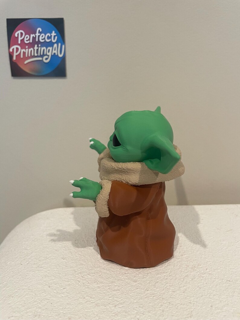 May include: A green figurine of a character with large ears and a brown robe. The figurine has white fingernails and is standing on a white surface. The figurine is a character from a popular science fiction franchise.