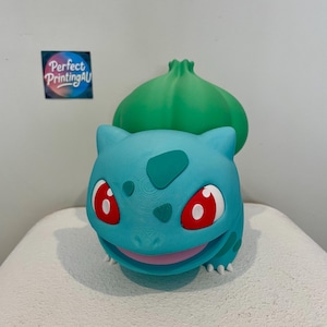 Puede incluir: Figura de Bulbasaur impresa en 3D en color turquesa, con un bulbo verde en la espalda. Los ojos rojos tienen reflejos blancos y una boca sonriente. La figura está sobre una superficie blanca.