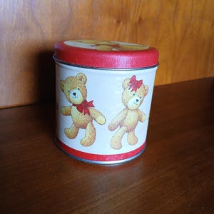 May include: A round metal tin with a red lid and a white body. The body features two cartoon teddy bears with red bows. The tin is sitting on a wooden surface.
