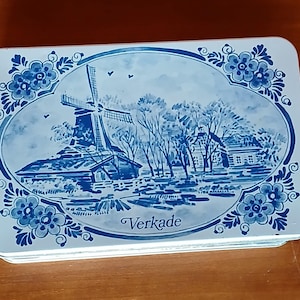 May include: A rectangular tin with a blue and white Dutch windmill scene. The design features a windmill, trees, and a house, framed by floral patterns. The word "Verkade" is printed on the tin.