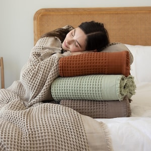 Peut inclure: Une pile de trois couvertures rectangulaires à tissage gaufré en orange rouille, vert sauge et taupe, chacune dotée de bords à franges. Une couverture beige à tissage gaufré est drapée sur la pile, présentant un motif en grille texturé.