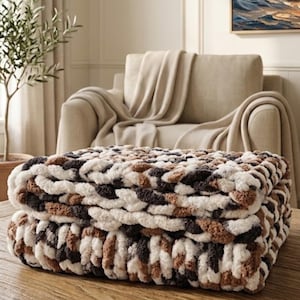 May include: A chunky knit throw blanket featuring a variegated pattern of thick, soft yarn in shades of cream, tan, and dark brown. The blanket is folded into a rectangular shape, showcasing the large, interlocking loops of the textured yarn.