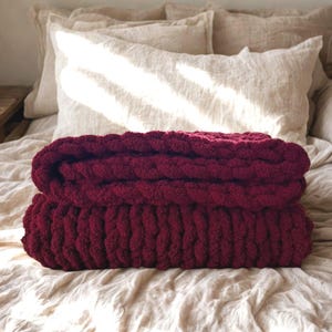 May include: A deep burgundy chunky knit blanket folded into two thick layers. The blanket features a prominent, textured loop stitch pattern created with thick yarn, showcasing a consistent, dense, and soft appearance throughout the entire piece.