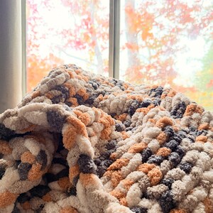 Cozy Toasted Birch chunky blanket draped near window with autumn colors outside, soft neutral tones of gray, cream, and tan.