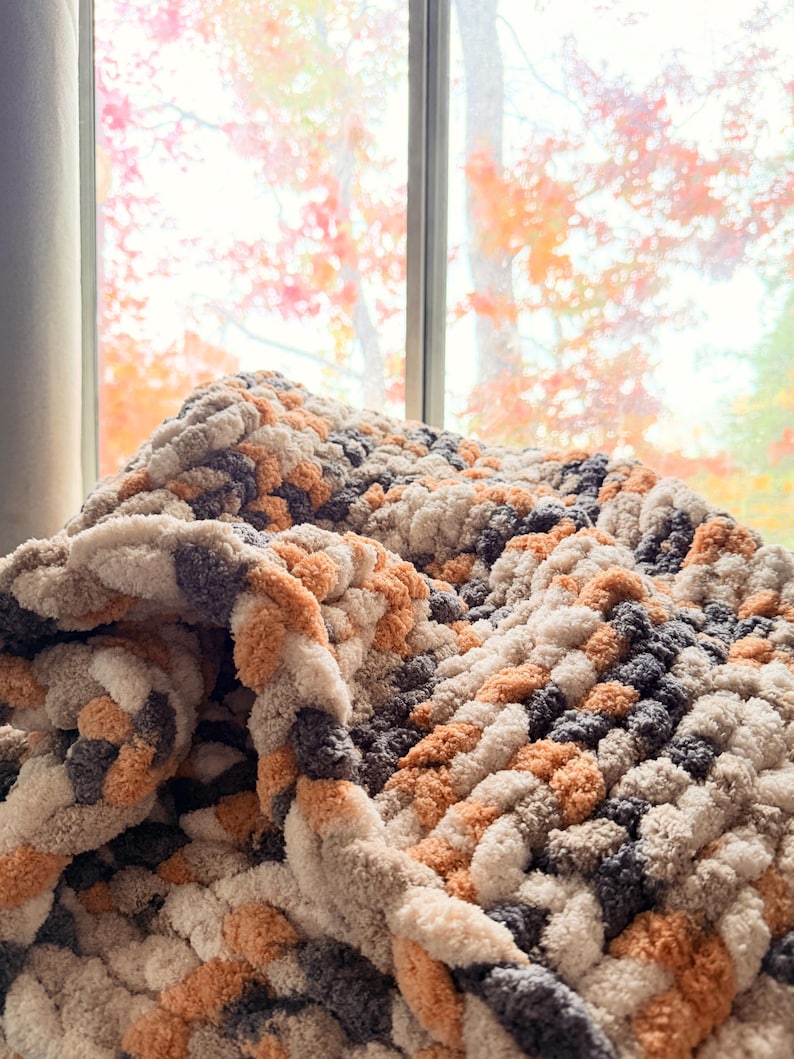 Cozy Toasted Birch chunky blanket draped near window with autumn colors outside, soft neutral tones of gray, cream, and tan.