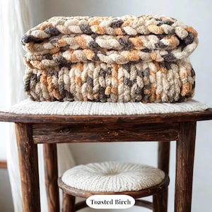 Hand-knit chunky blanket in Toasted Birch multi-tone yarn of cream, beige, and gray, folded on wood stool in natural light showing plush texture.
