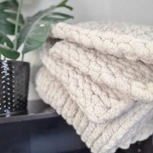 A folded chunky knit blanket in creamy French Vanilla beside green plant on dark table, showing soft texture and neutral home style.