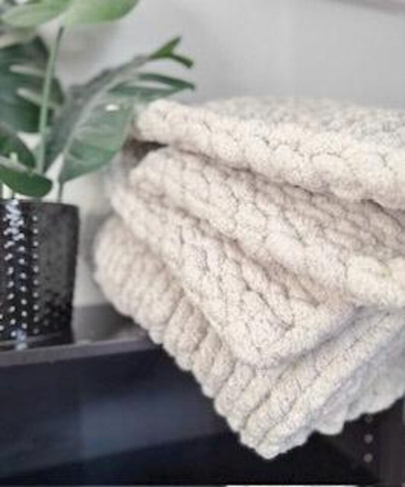 A folded chunky knit blanket in creamy French Vanilla beside green plant on dark table, showing soft texture and neutral home style.