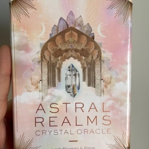 May include: A rectangular box for the "Astral Realms Crystal Oracle" deck. The box features a pastel color scheme with an arched structure, crystals, and the moon. The text "Astral Realms Crystal Oracle" is displayed in a copper color.