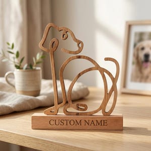 Op de afbeelding: Een lichtbruin houten sculptuur in de vorm van een doorlopende lijntekening van een zittende hond. Het beeldhouwwerk is gemonteerd op een rechthoekige houten voet met de tekst CUSTOM NAME in hoofdletters gegraveerd op de voorzijde.