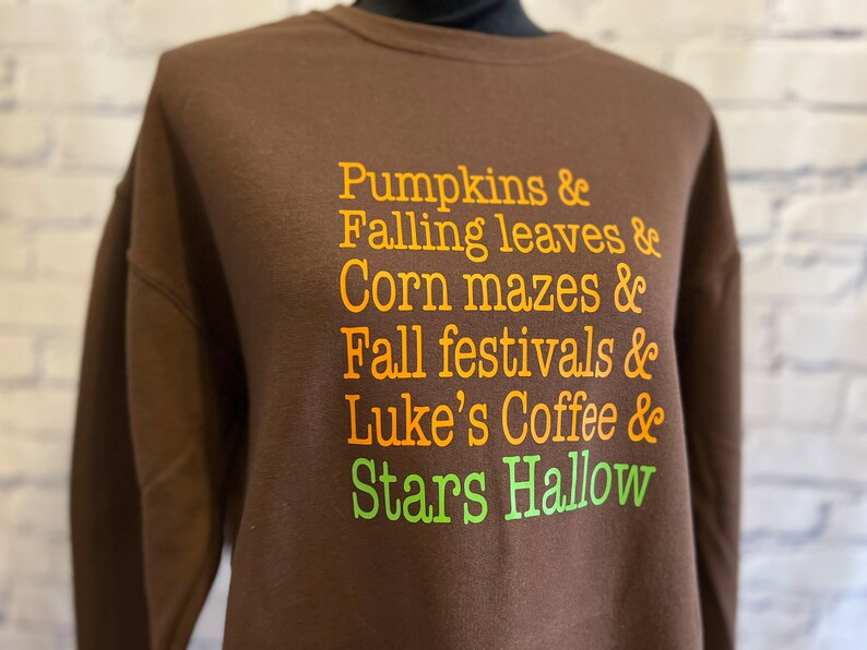 May include: Brown sweatshirt with orange text that reads "Pumpkins & Falling leaves & Corn mazes & Fall festivals & Luke's Coffee & Stars Hallow"