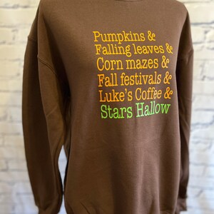 May include: Brown sweatshirt with a white crew neck and a graphic print of the text "Pumpkins & Falling leaves & Corn mazes & Fall festivals & Luke's Coffee & Stars Hallow" in orange, yellow, and green.