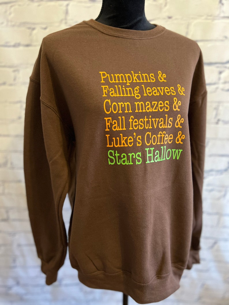 May include: Brown sweatshirt with a white crew neck and a graphic print of the text "Pumpkins & Falling leaves & Corn mazes & Fall festivals & Luke's Coffee & Stars Hallow" in orange, yellow, and green.