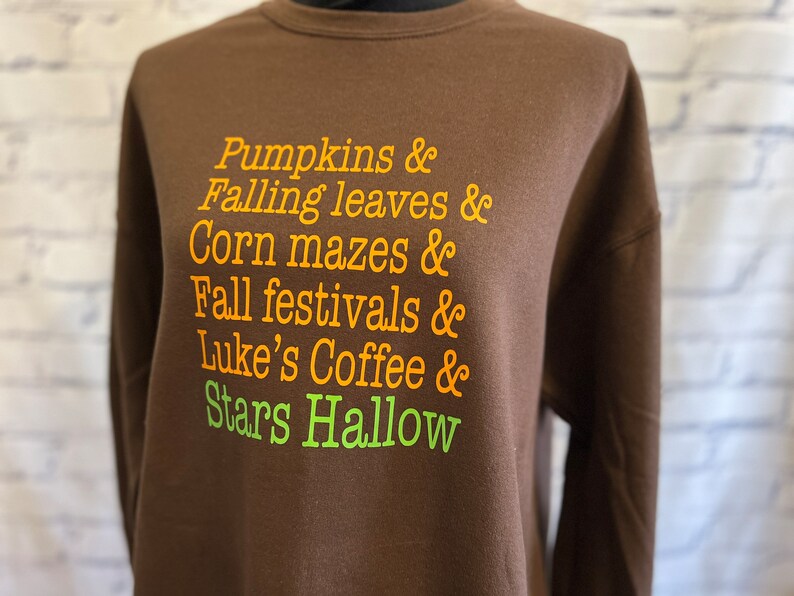 May include: Brown sweatshirt with orange and green text that reads "Pumpkins & Falling leaves & Corn mazes & Fall festivals & Luke's Coffee & Stars Hallow"