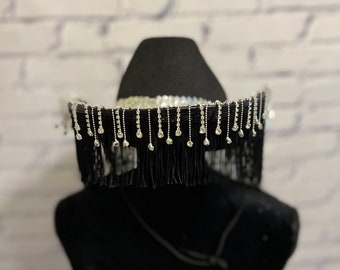 May include: A black cowboy hat with a fringe trim and rhinestones. The hat is on a black mannequin head.