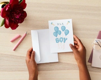 Peut inclure: Une carte de vœux avec le texte "It's a Boy" et des graphiques de ballons bleus est tenue ouverte à côté d'une enveloppe blanche. Un stylo rose et une fleur rouge sont également visibles.