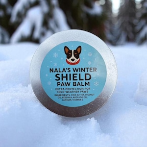 May include: A round, silver tin of Nala's Winter Shield Paw Balm, designed for extra protection for cold weather paws. The label features a cartoon dog illustration and snowflakes on a light blue background. Ingredients include shea butter, coconut oil, beeswax, avocado oil, lanolin, and vitamin E.