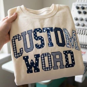 May include: A cream-colored crewneck sweatshirt featuring the text CUSTOM WORD in large, block-style letters. Each letter is filled with a different navy blue and white patterned fabric, including gingham, polka dots, stripes, and floral prints.