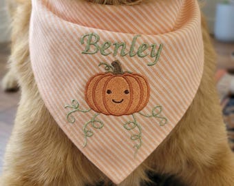 May include: A light orange and white striped pet bandana featuring an embroidered orange pumpkin with a smiling face and green vines. The name Benley is embroidered in green script lettering above the pumpkin design on the fabric.