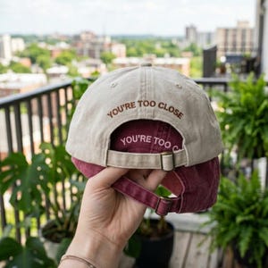 May include: A beige baseball cap with the text "YOU'RE TOO CLOSE" embroidered in brown on the back. The underside of the brim is burgundy, and the adjustable strap is also burgundy.