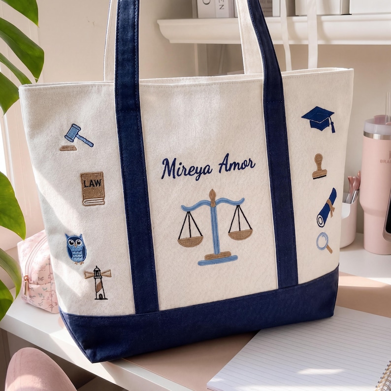 K&ouml;nnte beinhalten: Cremefarbene Tragetasche mit marineblauen Akzenten und Griffen. Die Tasche zeigt gestickte Symbole zum Thema Recht, den Namen "Mireya Amor" und eine Waage. Der Boden und die Verzierungen sind marineblau.