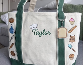 May include: Cream canvas tote bag with green trim and handles. The bag is decorated with embroidered baking-themed images and the name "Taylor". A bread-shaped keyring hangs from the handle. Designed for carrying items.