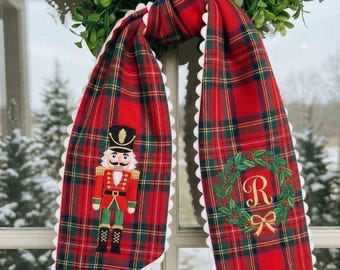 May include: A red, green, and yellow tartan fabric scarf featuring a white pom-pom trim. One end displays an embroidered nutcracker in a red uniform, while the other end features an embroidered green wreath with a gold bow and the letter R.
