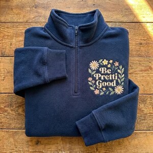 May include: A navy blue fleece pullover with a quarter-zip neckline and ribbed cuffs. The pullover features an embroidered floral design with the words "Be Pretti Good" in the center. The fleece is soft and appears comfortable.