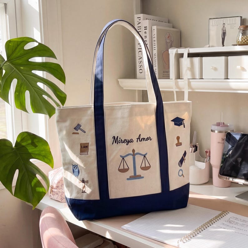 K&ouml;nnte beinhalten: Cremefarbene Tragetasche mit marineblauen Akzenten und Griffen. Die Tasche zeigt gestickte Symbole zum Thema Recht und den Text "Mireya Amor" &uuml;ber einer Waage der Gerechtigkeit. Ein stilvolles und praktisches Accessoire.
