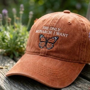 May include: A rust-colored baseball cap with the embroidered text "THE ONLY MONARCH I WANT" above a butterfly design. The butterfly is orange, black, and white. The cap is made of a soft fabric and has a curved brim.