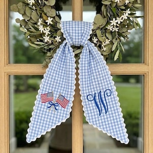 May include: A blue and white gingham door bow with a scalloped edge and white trim. The bow features embroidered American flags and a blue monogram letter "W". A green wreath with white flowers is in the background.