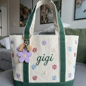 Puede incluir: Bolso tote color crema con detalles y asas verdes, bordado con flores coloridas. La palabra "gigi" est&aacute; bordada en verde. Un charm de flor morada y un peque&ntilde;o pomp&oacute;n cuelgan del asa.