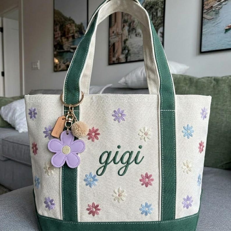 Puede incluir: Bolso tote color crema con detalles y asas verdes, bordado con flores coloridas. La palabra "gigi" est&aacute; bordada en verde. Un charm de flor morada y un peque&ntilde;o pomp&oacute;n cuelgan del asa.
