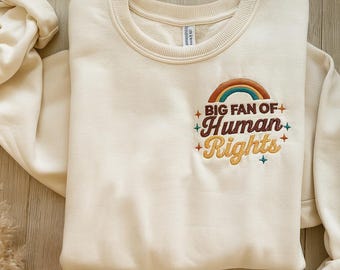 May include: Cream-colored sweatshirt with a crew neck and long sleeves. The front features an embroidered rainbow and the text "BIG FAN OF+ Human Rights+" in a mix of colors. The sweatshirt is folded on a wooden surface.