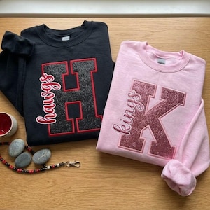 May include: A black crewneck sweatshirt featuring a large glittery letter H with the word hawgs in script, and a light pink crewneck sweatshirt featuring a large glittery letter K with the word kings in script, both with long sleeves.