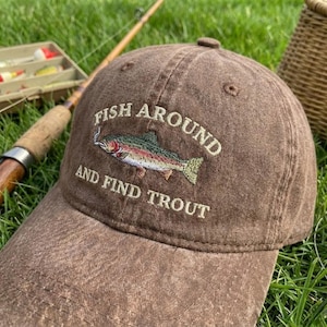 May include: Brown baseball cap with the embroidered text "FISH AROUND AND FIND TROUT" and a colorful trout illustration. The cap is made of a textured fabric and has a curved brim. Fishing gear is visible in the background.