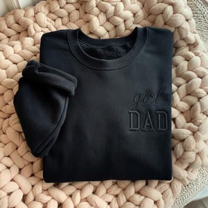May include: Black sweatshirt with the embroidered text "girl DAD". The sweatshirt has a crew neck and long sleeves. The sleeve is folded up. The sweatshirt is displayed on a beige knitted blanket.
