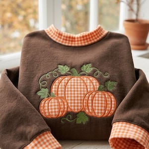 May include: A brown long-sleeve sweatshirt featuring an embroidered design of three pumpkins with an orange and white gingham pattern. The collar and sleeve cuffs are trimmed with a matching orange and white gingham fabric, and the pumpkins are accented with green embroidered leaves and vines.