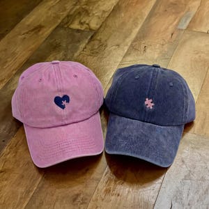 May include: Two baseball caps, one pink and one navy blue, are displayed. The pink cap features a dark blue heart-shaped puzzle piece, while the navy cap has a pink puzzle piece. Both caps have a distressed, vintage look.