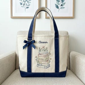 May include: Cream tote bag with navy blue trim and handles. It features a stack of books with floral embroidery and the name "Eleanor". A navy blue bow is attached to the handle. The bag is on a beige chair.