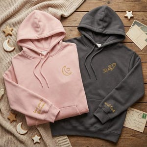 May include: Two hooded sweatshirts, one pink and one charcoal gray, are displayed on a wooden surface. The pink hoodie has a crescent moon and heart design, with "Love you to the moon" on the sleeve. The gray hoodie features a rocket and heart design, with "and back" on the sleeve.