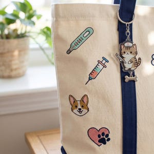 May include: Cream tote bag with navy trim and a keychain. The bag has embroidered images of a thermometer, syringe, Corgi dog, paw print, and a cat wearing a nurse's hat. The keychain has a cat charm and a bone charm.
