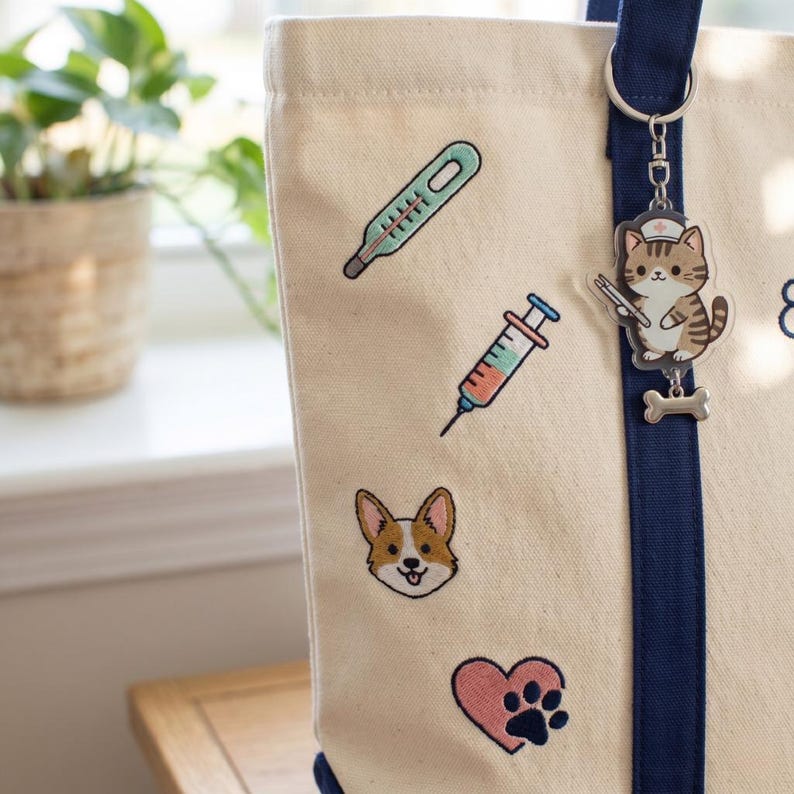 May include: Cream tote bag with navy trim and a keychain. The bag has embroidered images of a thermometer, syringe, Corgi dog, paw print, and a cat wearing a nurse's hat. The keychain has a cat charm and a bone charm.