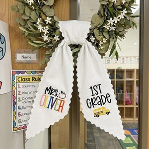May include: A green eucalyptus wreath with white flowers features a white fabric bow with scalloped edges. The left tail displays the text "MRS. OLIVER" in colorful letters with an apple icon, and the right tail shows "1ST GRADE" with a yellow school bus.